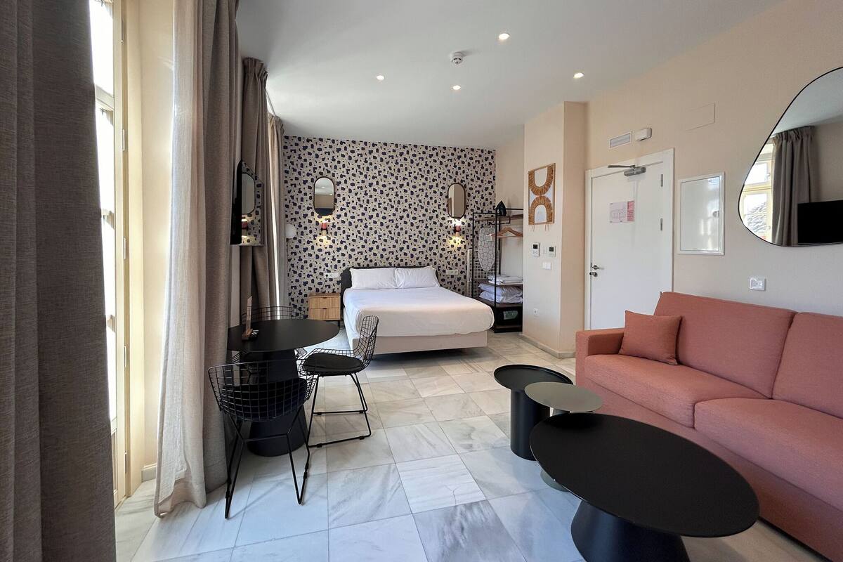 Una habitación de hotel moderna con cama, sofá, mesa de comedor con sillas, televisor y pared con diseño.