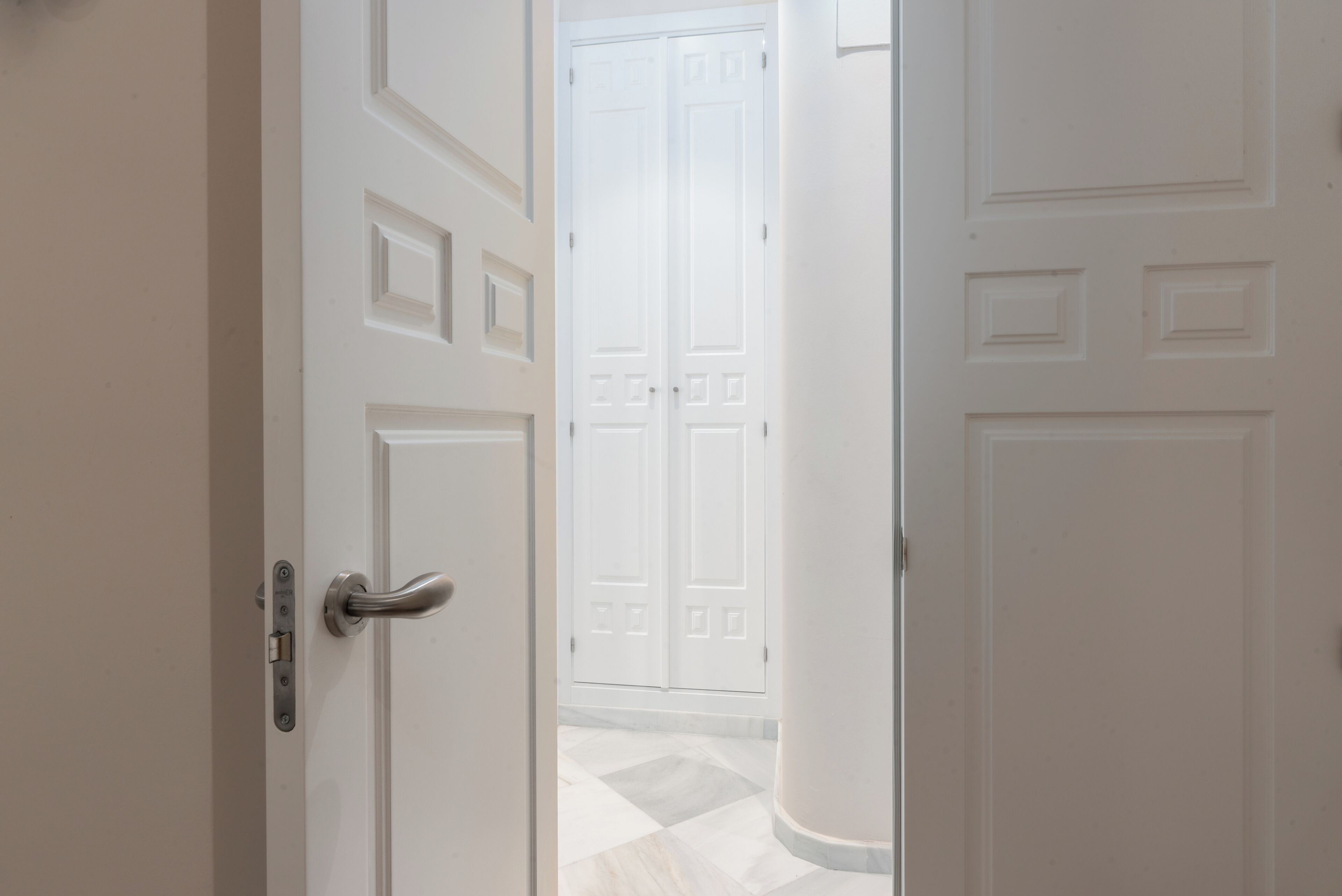 A white door with a silver handle and a lock, leading to a room with a tiled floor and a white door in the background.