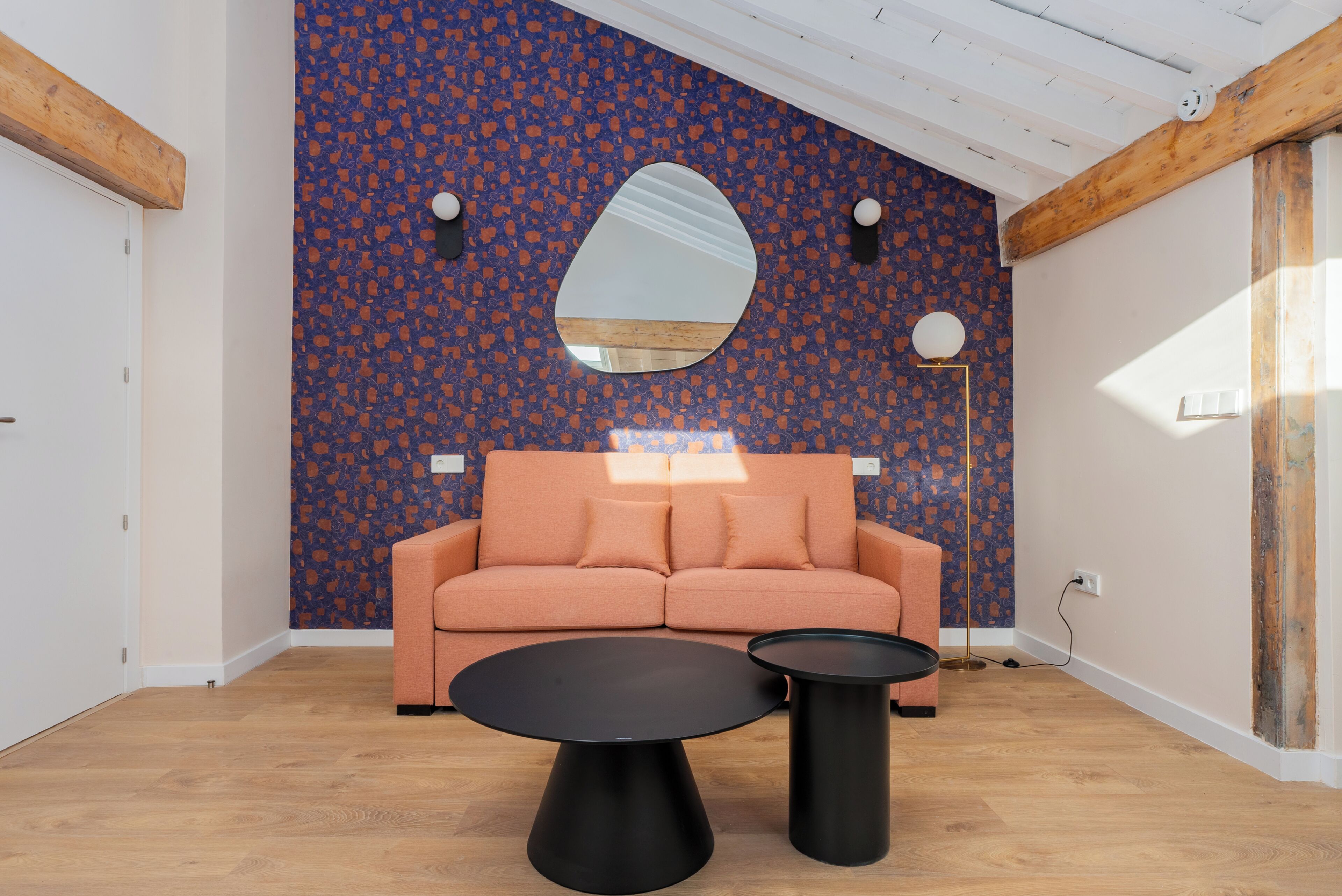 Una sala de estar moderna con una pared estampada, un sofá, dos mesas negras y un espejo grande.