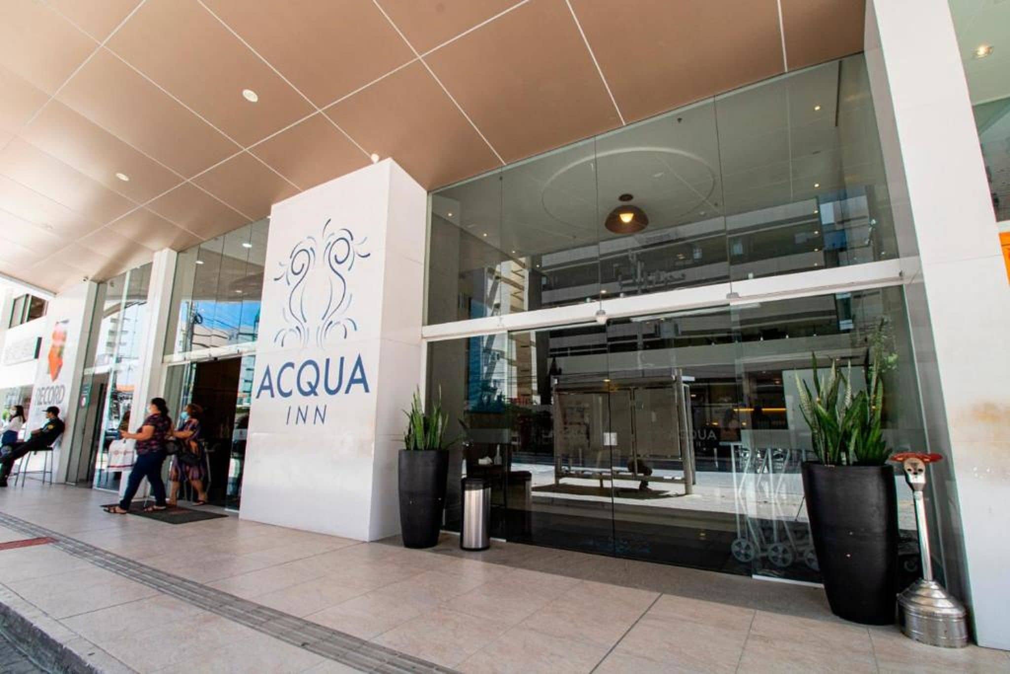 The exterior of the ACQUA INN, featuring a modern glass facade, a prominent logo, and potted plants.