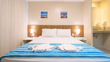 Habitación de hotel con cama, dos mesitas de noche, un tocador, baño con lavamanos y espejo, y dos cuadros en la pared.
