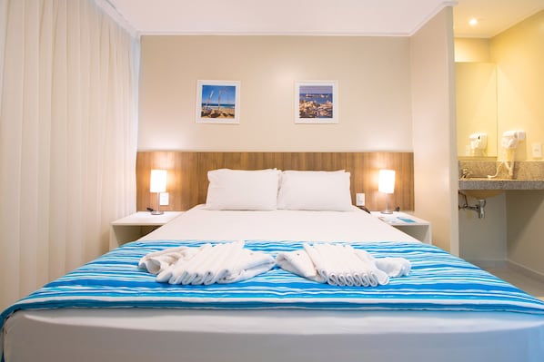 A hotel room with a bed, two nightstands, a bedside table, a bathroom with a sink and mirror, and two framed pictures on the wall. - Acqua Inn Maceio Ponta Verde (Maceió)