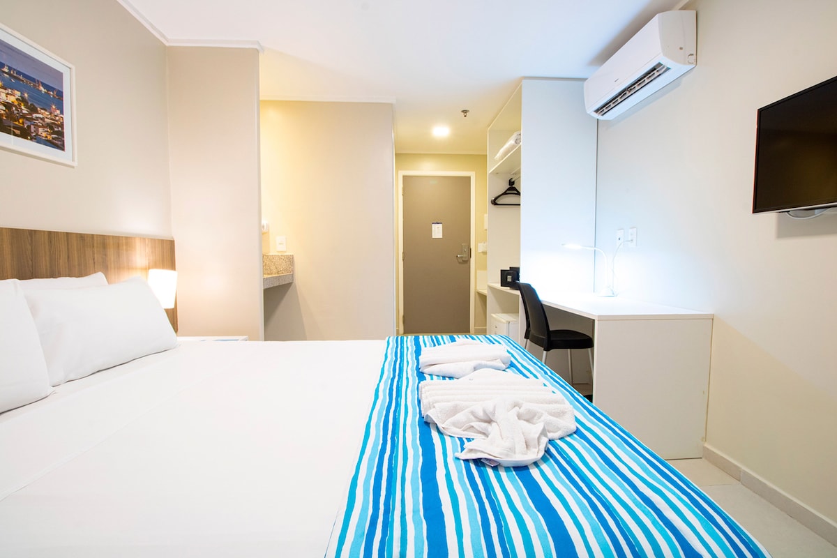 A hotel room with a bed, a desk, a chair, a TV, and an air conditioner.