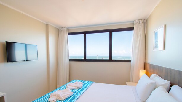 A hotel room with a large bed, a flat-screen TV mounted on the wall, a window with a view of the sea, and a bedside table with a lamp. - Acqua Inn Maceio Ponta Verde (Maceió)