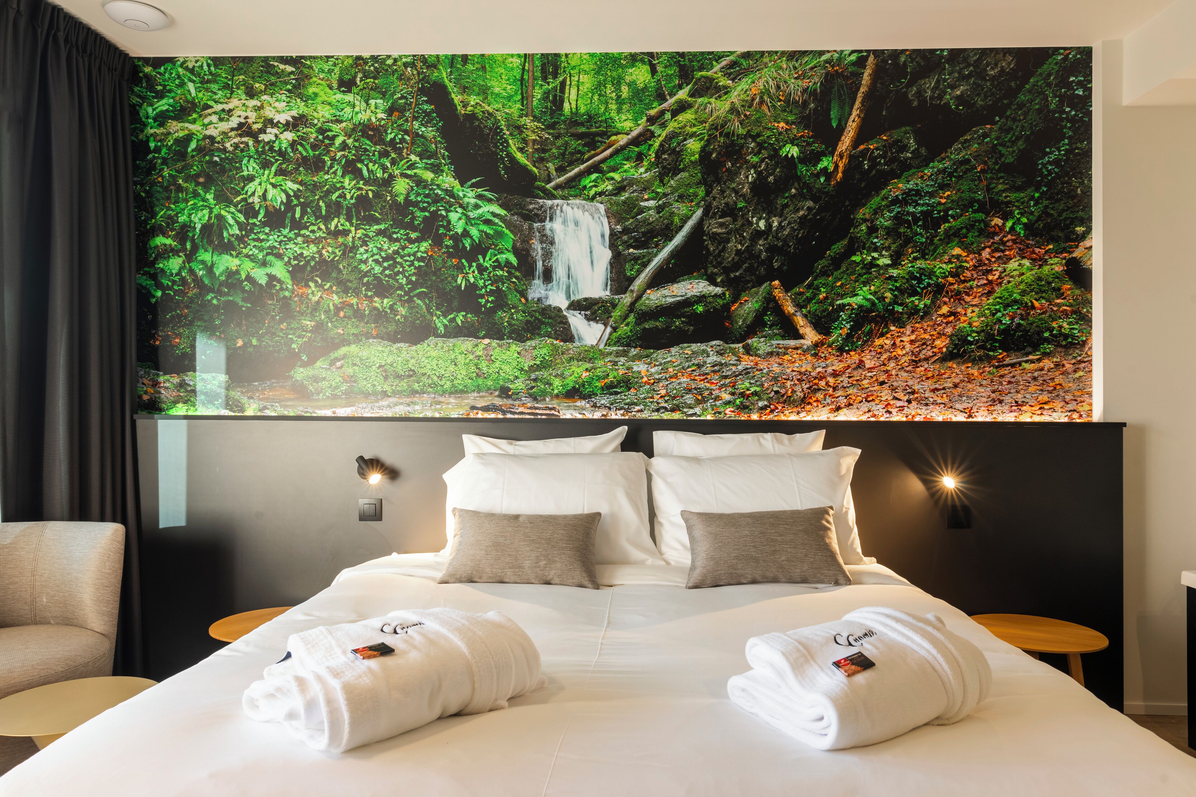Ein Hotelzimmer mit einem großen Bett, zwei Nachttischen, einem am Wand montierten Fernseher, der eine Waldszene zeigt, und einem dunkel gefärbten Kopfteil.