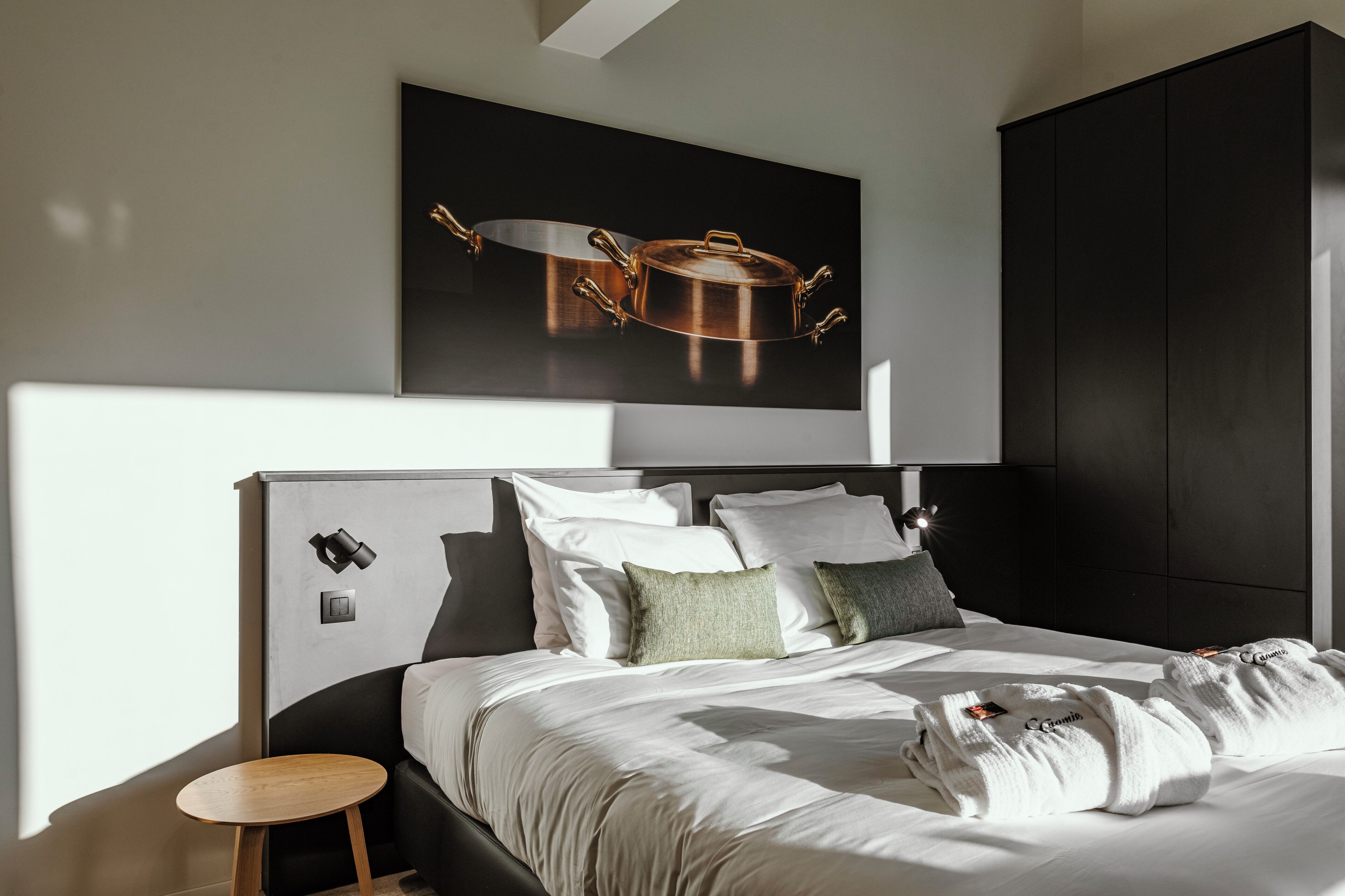Une chambre d’hôtel moderne avec un grand lit, une petite table d’appoint en bois, une œuvre d’art représentant des ustensiles de cuisine accrochée au mur et une armoire sombre.