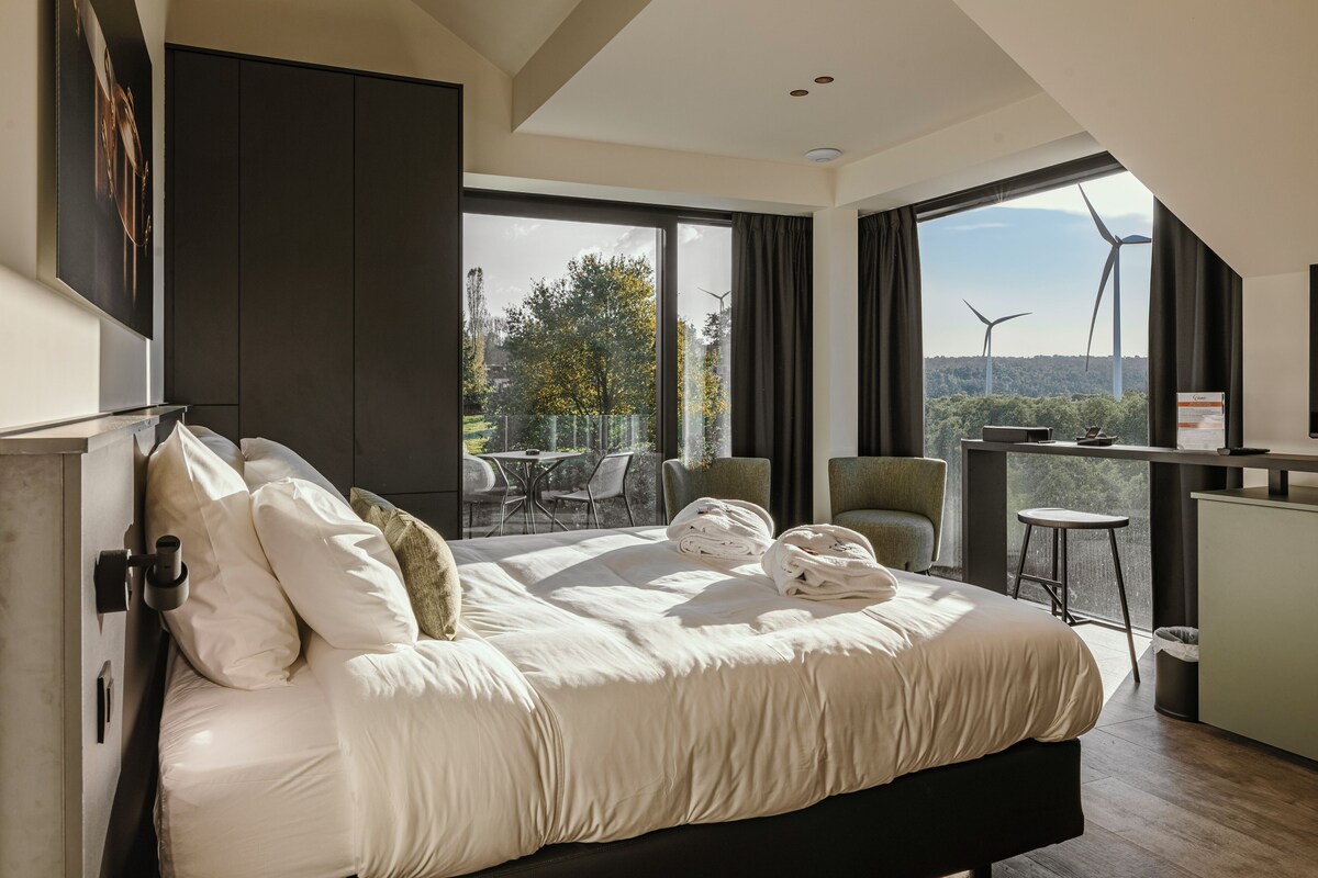 A modern hotel room with a large bed, a balcony with seating, and a view of wind turbines.