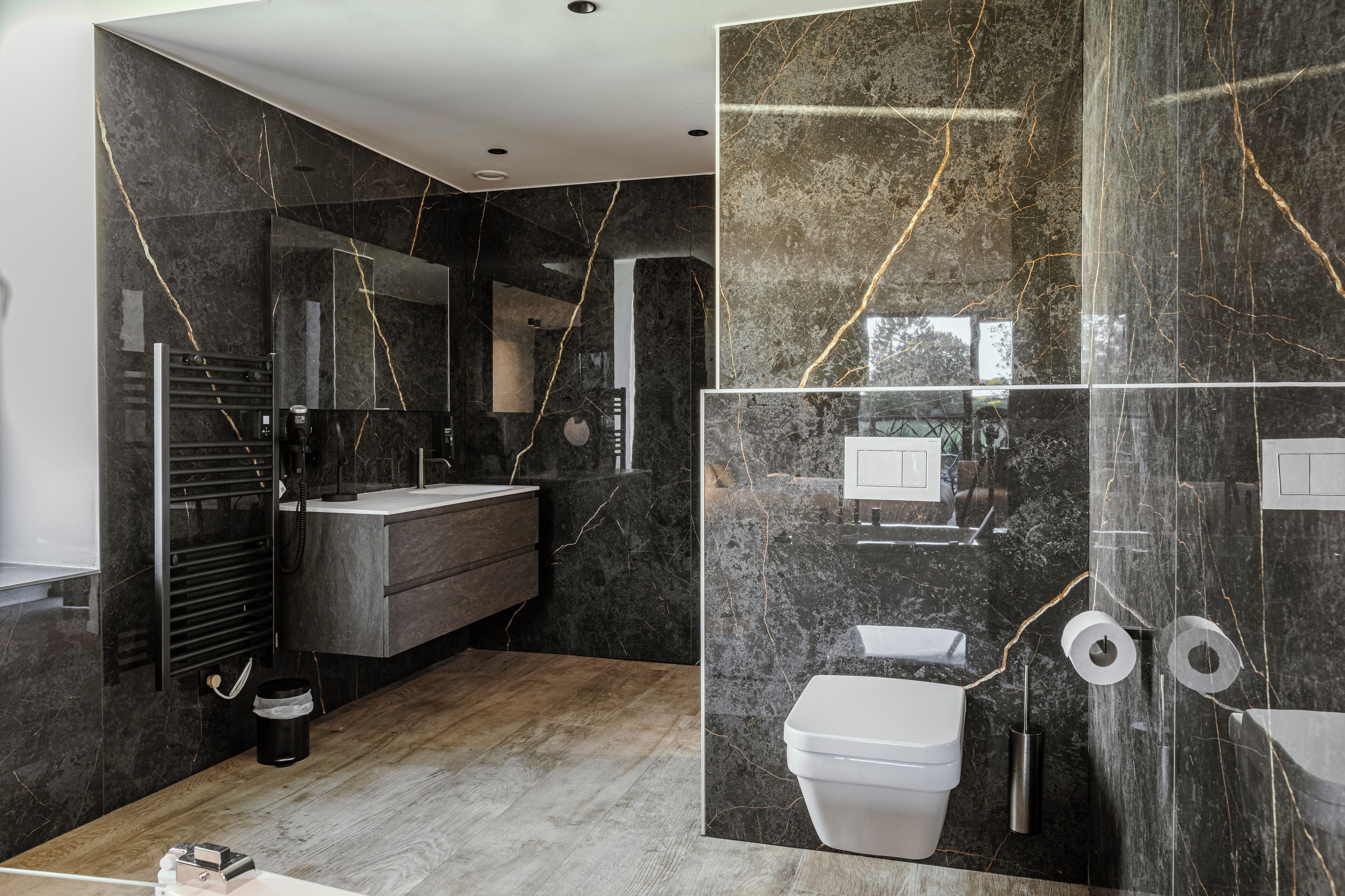 A modern bathroom with dark marble walls, a wooden floor, a white toilet, and a black towel rack.
