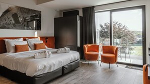 A modern hotel room with a large bed, a seating area with two chairs, a large window with a view, and a painting on the wall. - CC Nomie Hôtel Restaurant (Dinant)
