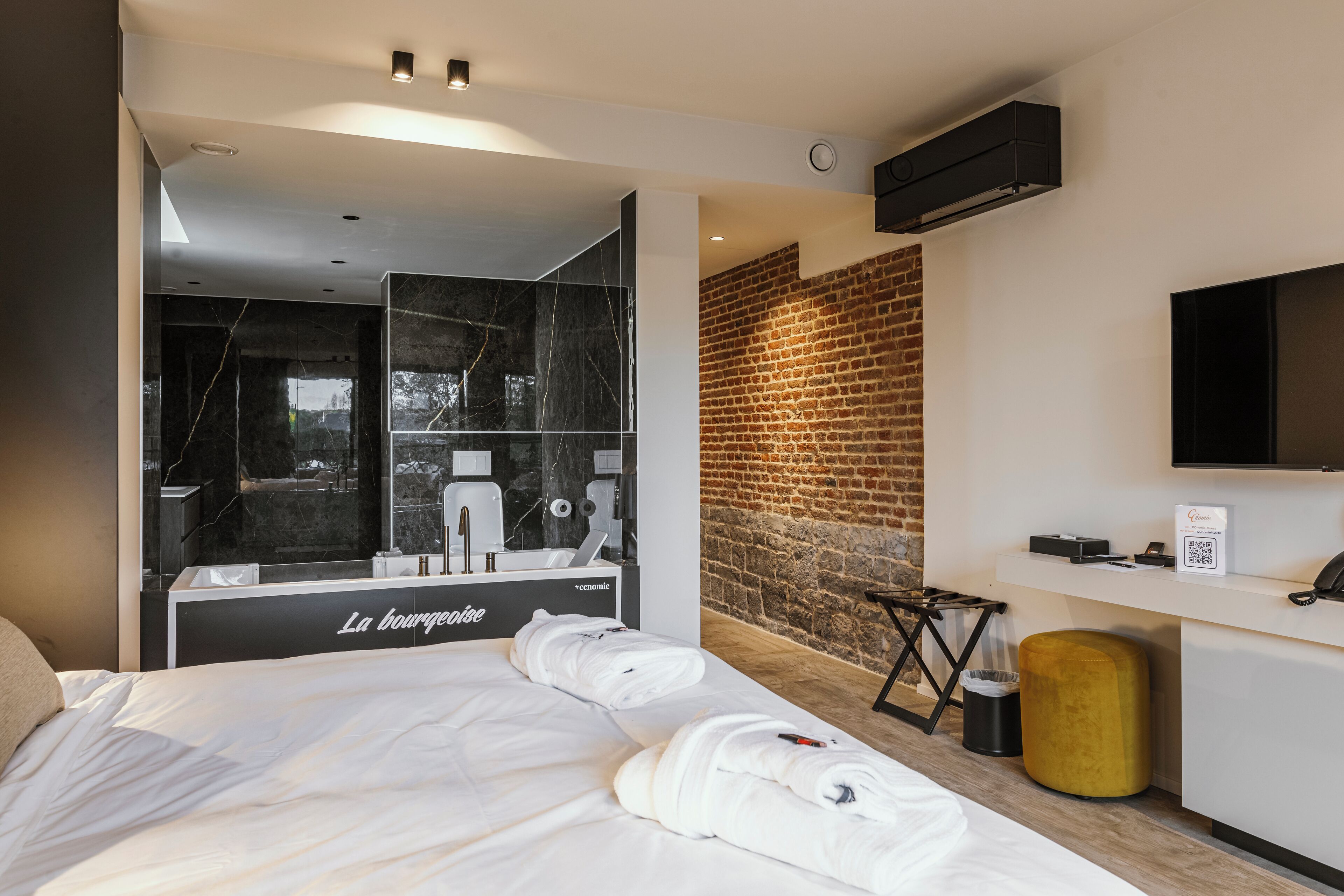 A modern hotel room with a large bed, a mirror, a brick wall, a TV, and a minibar.