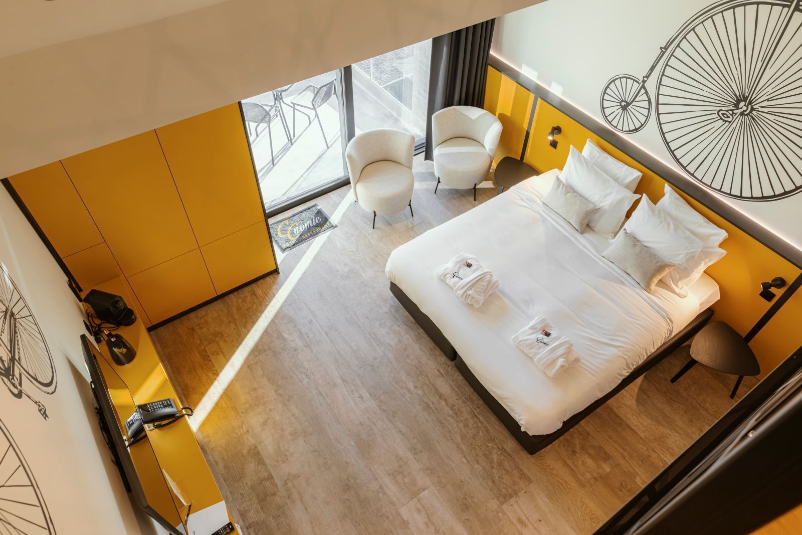 Una habitación de hotel moderna con una cama grande, dos sillas, un escritorio con una computadora y una obra de arte con motivo de bicicleta.