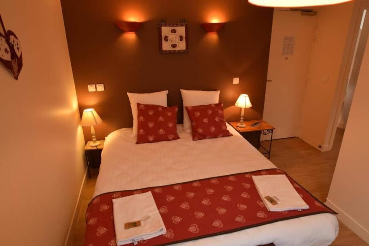 Ein Hotelzimmer mit einem großen Bett, zwei Nachttischlampen, einer roten, gemusterten Bettdecke und einem kleinen Tisch mit einer Lampe.