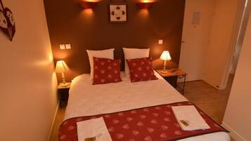 A hotel room with a large bed, two bedside lamps, a red patterned bedspread, and a small table with a lamp.