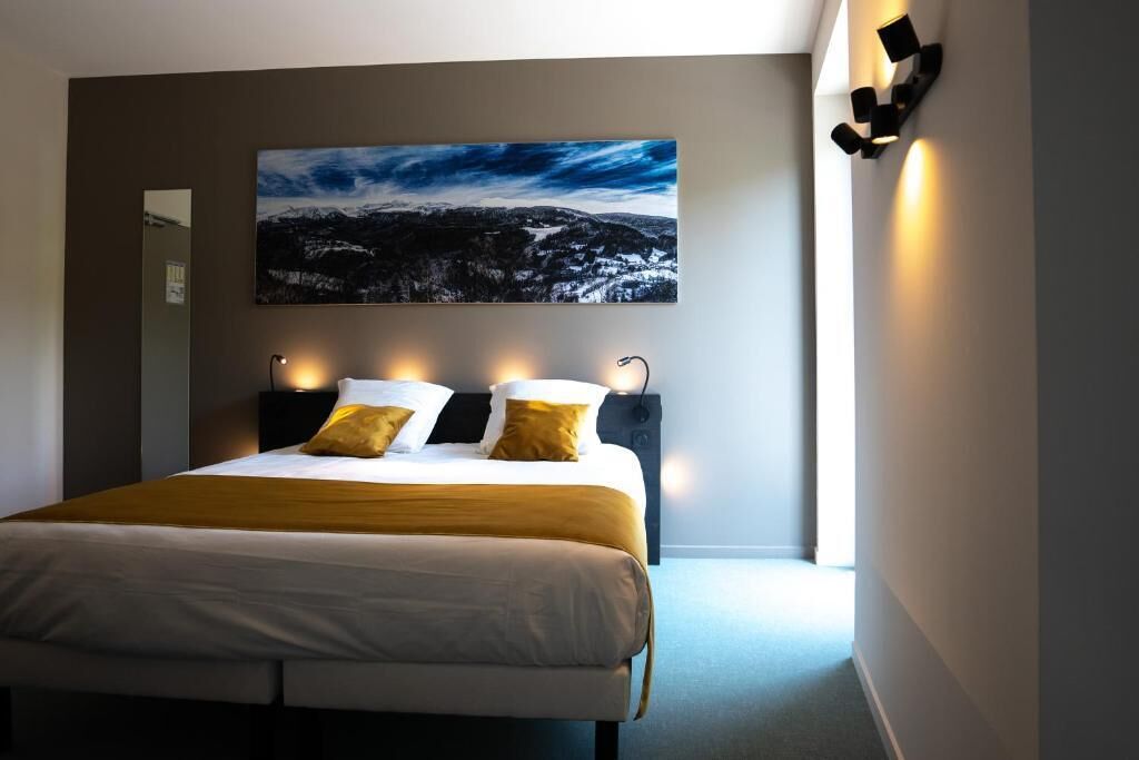 A bedroom with a large bed, a nightstand, a wall-mounted lamp, and a painting of a snowy landscape.