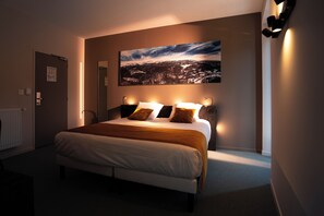 A hotel room with a large bed, a painting on the wall, and a view of a cityscape. - Hôtel Le Genève (La Bourboule)