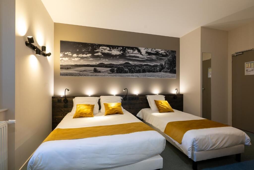 A hotel room with two beds, each with white bedding and a yellow throw. There is a large black and white landscape painting above the beds, and wall-mounted lights above each bed.