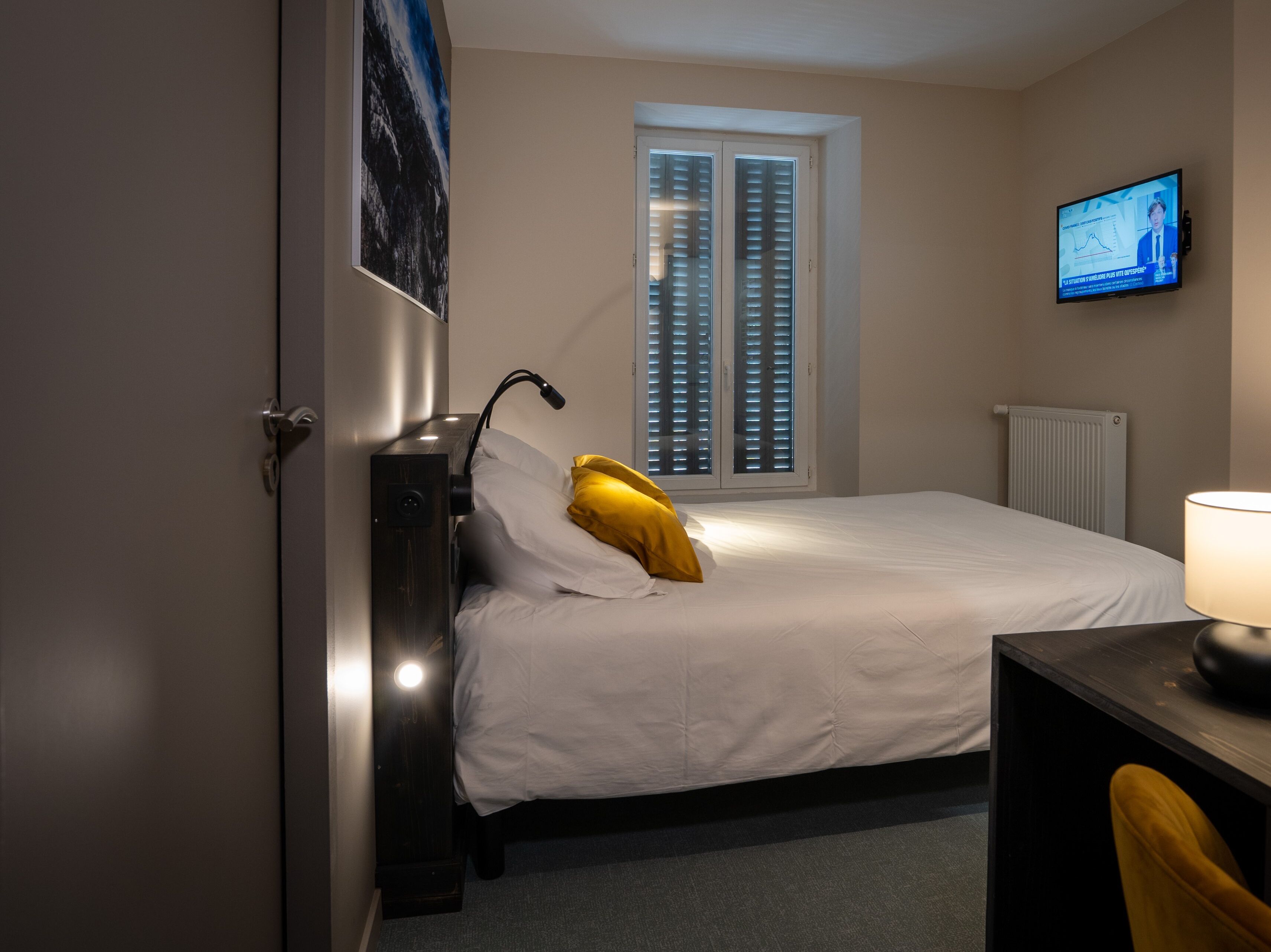A hotel room with a bed, a desk, a chair, a TV on the wall, and a window with blinds.