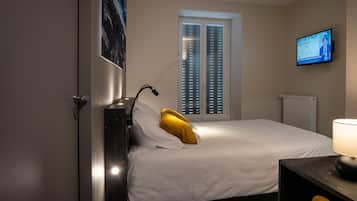 A hotel room with a bed, a desk, a chair, a TV on the wall, and a window with blinds.