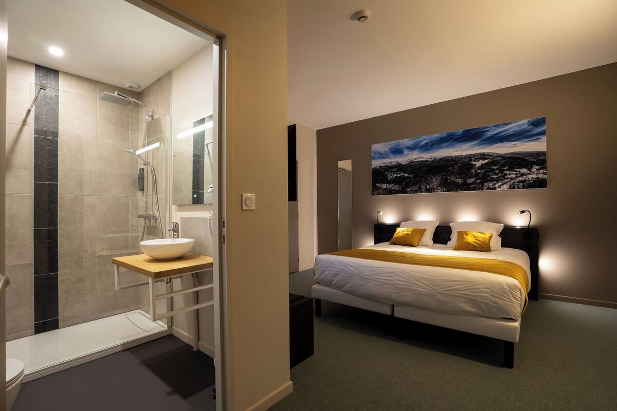 Ein modernes Hotelzimmer mit bodenebenen Dusche, Nachttisch, Bett mit Kopfteil und großem Landschaftsbild an der Wand.