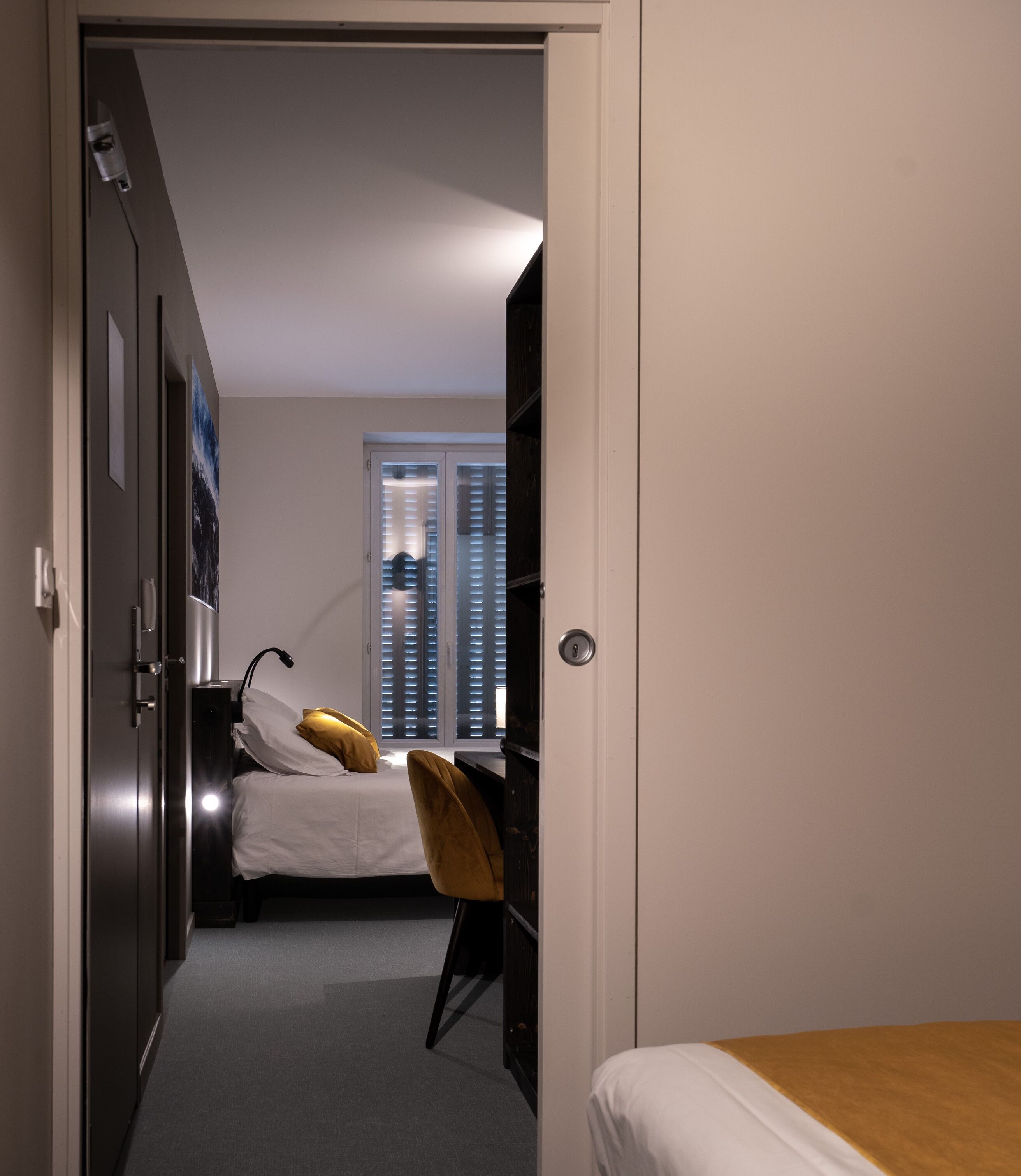 A bedroom with a bed, a desk, and a chair. There is a wardrobe and a window with blinds.