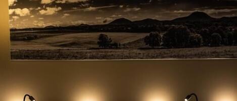 A bed with white linens and two pillows, a wall-mounted light fixture above, and a landscape painting depicting a cloudy sky and mountains.