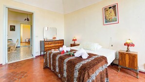 Habitación de hotel con cama, dos mesitas de noche, armario, espejo, un cuadro en la pared y un pasillo que da a otra habitación.