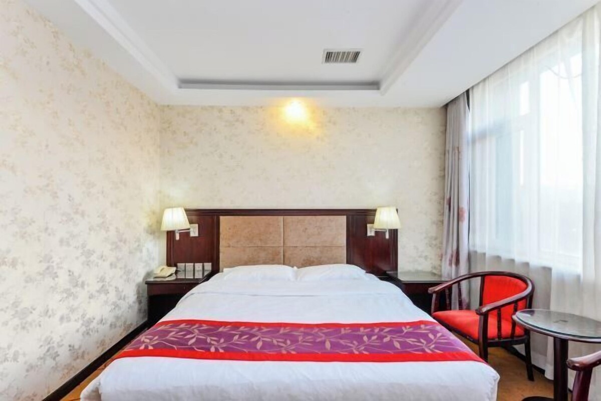 A hotel room with a bed, two bedside lamps, a small table, a chair, and a window with curtains.