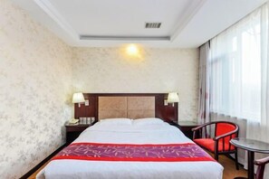 A hotel room with a bed, two bedside lamps, a small table, a chair, and a window with curtains.