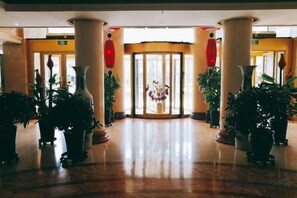 Un vestíbulo de hotel con piso de mármol, amplias columnas, plantas en macetas y una entrada con puerta de vidrio.