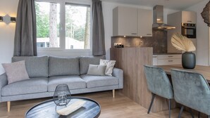 A modern living room with a grey sofa, a round coffee table, and a dining area with two chairs. A kitchen with a wooden island and built-in appliances is visible in the background. - Chalet in Eindhoven Near Green Forests (Mierlo)