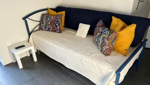 Un lit avec un drap blanc, deux oreillers colorés, une table de chevet blanche avec une télécommande, et une structure en métal bleu foncé.