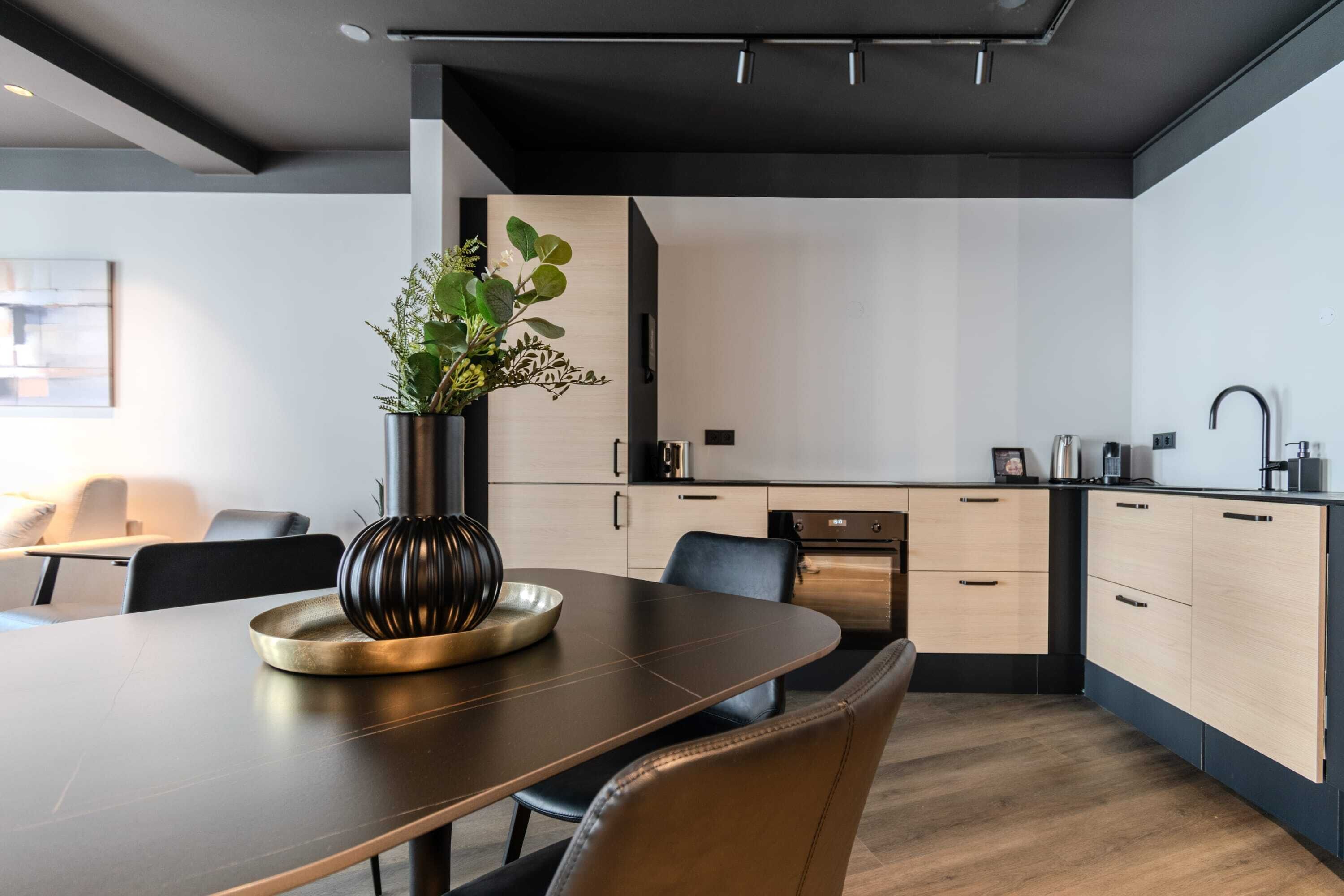 Una cocina moderna con zona de comedor, que incluye una mesa de madera oscura, sillas negras, un florero con follaje y electrodomésticos de acero inoxidable.