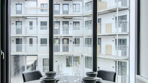 Comedor con mesa y sillas, ventanas amplias y vista a un edificio de varios pisos.