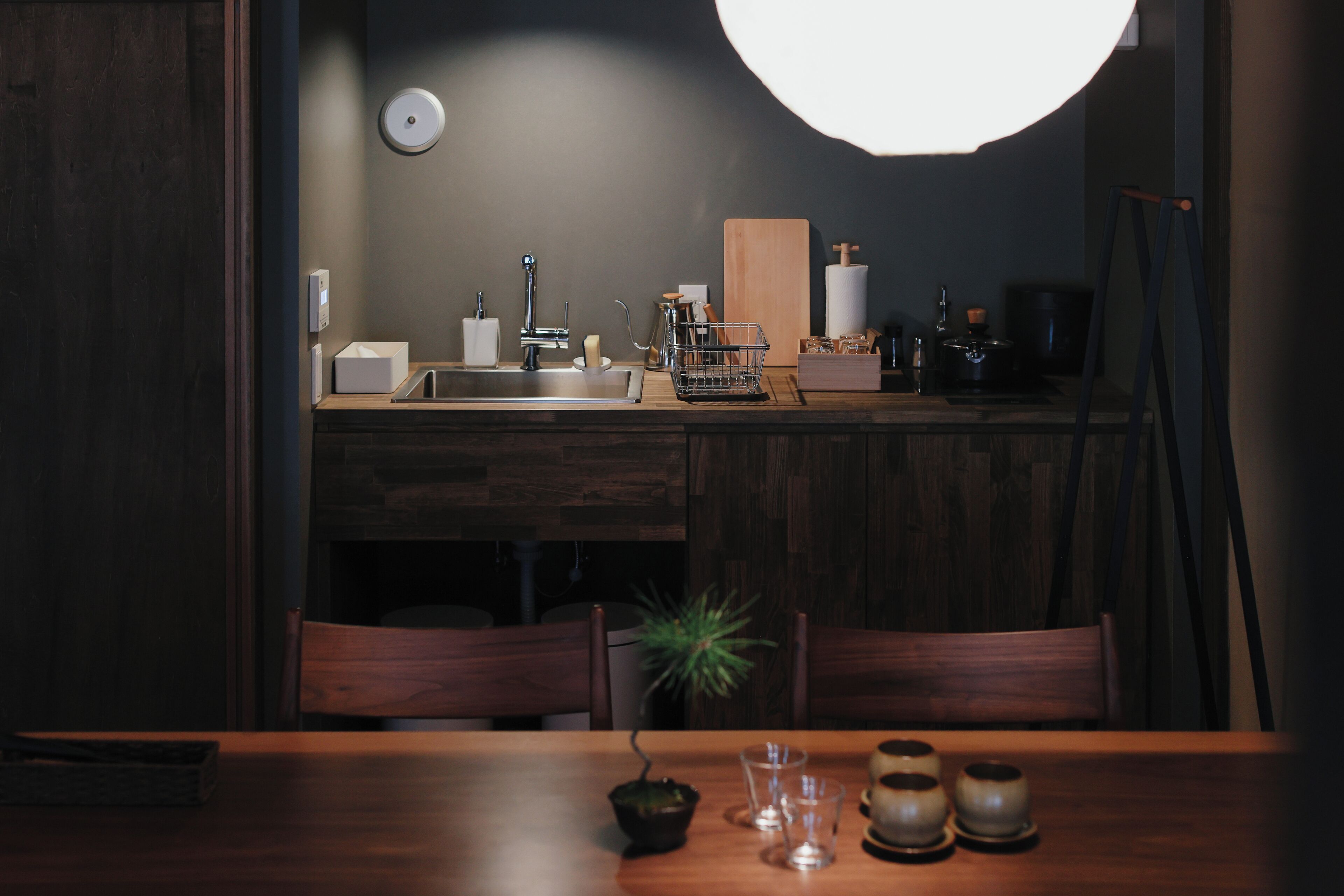 A dimly lit kitchen with a wooden table, chairs, and a potted plant.