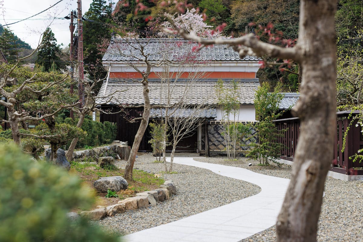 Un edificio japonés tradicional con tejado de tejas, rodeado por un camino de grava y pequeños árboles.