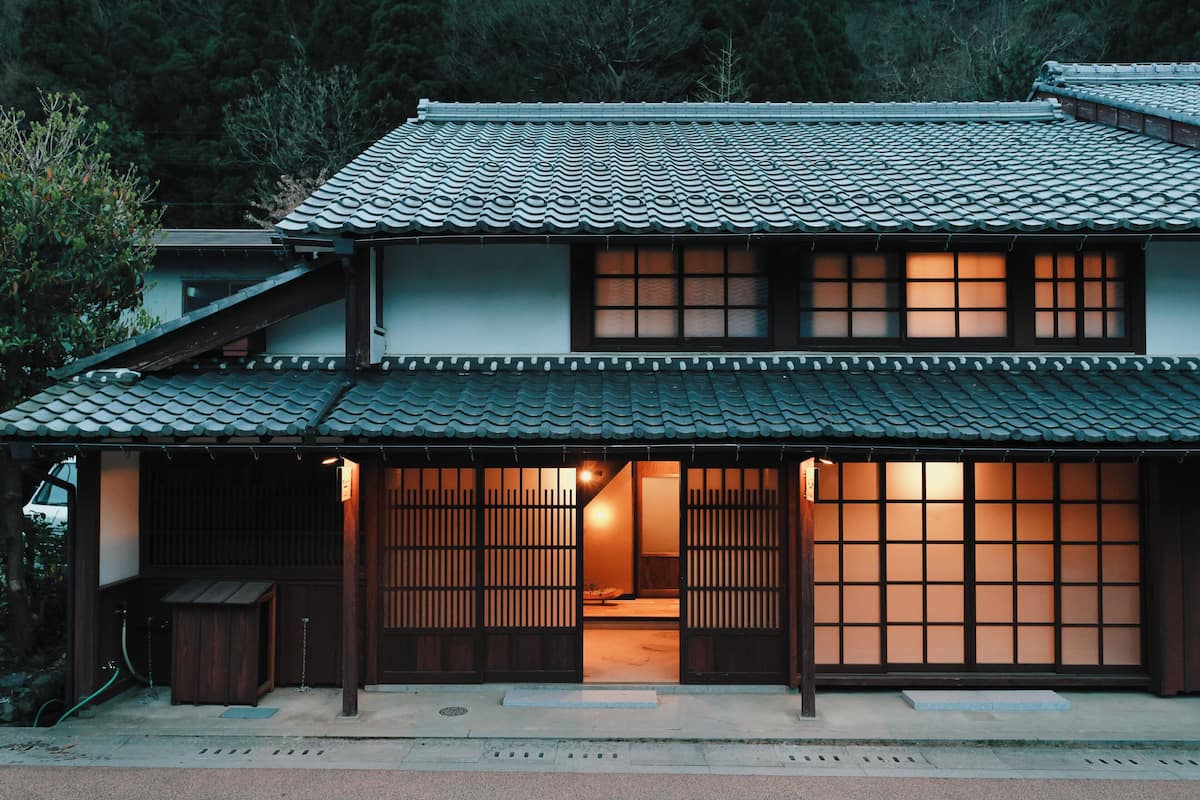 Una casa tradicional japonesa con tejado de tejas, puertas correderas y un interior visible con una fuente de luz.