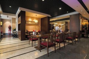 Een moderne hotellobby met een eetgedeelte, inclusief een lange houten tafel met stoelen, een receptiebalie en een decoratieve wand met een kroonluchter.