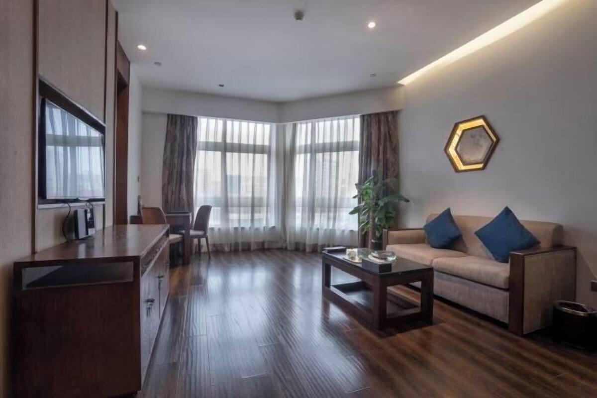 Una sala moderna con sofá, mesa de centro y televisor sobre un mueble de madera. Amplias ventanas con cortinas y una planta en el alféizar.