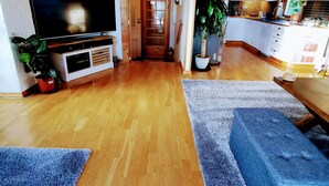 Amplia sala de estar con suelo de madera, televisor de pantalla plana, una escalera, cocina con armarios blancos y una planta en maceta.