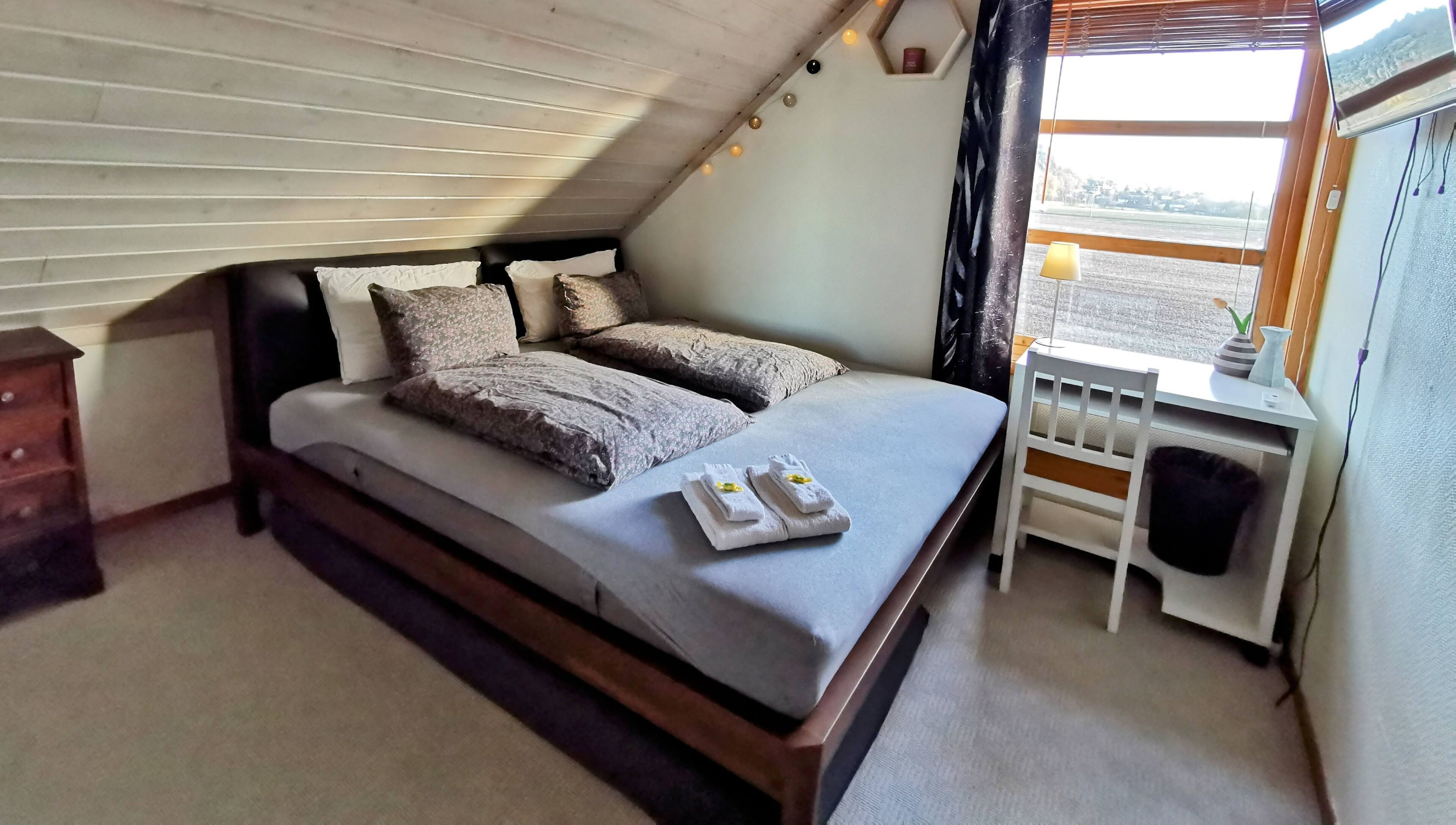 A neatly made bed with pillows and towels, a small bedside table with a lamp, and a view of the outside through a window.