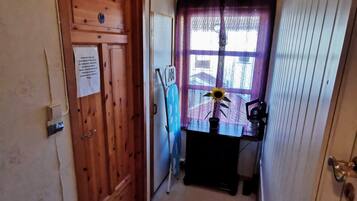 A narrow hallway with a wooden door on the left, a small table with a potted plant and a fan on the right, and a window with purple curtains.