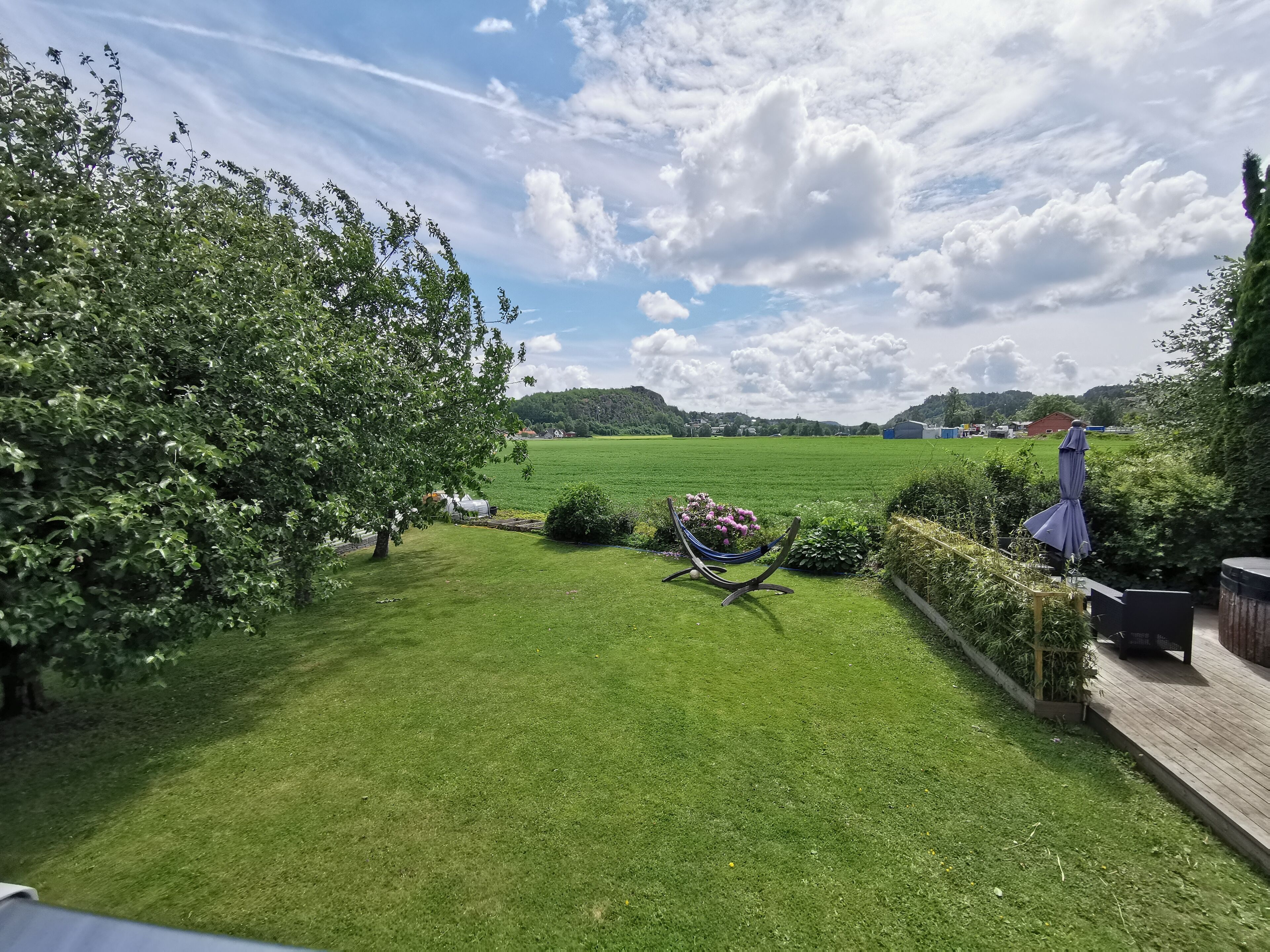 A well-maintained garden with a hammock, a wooden deck, and a view of a green field and distant buildings.
