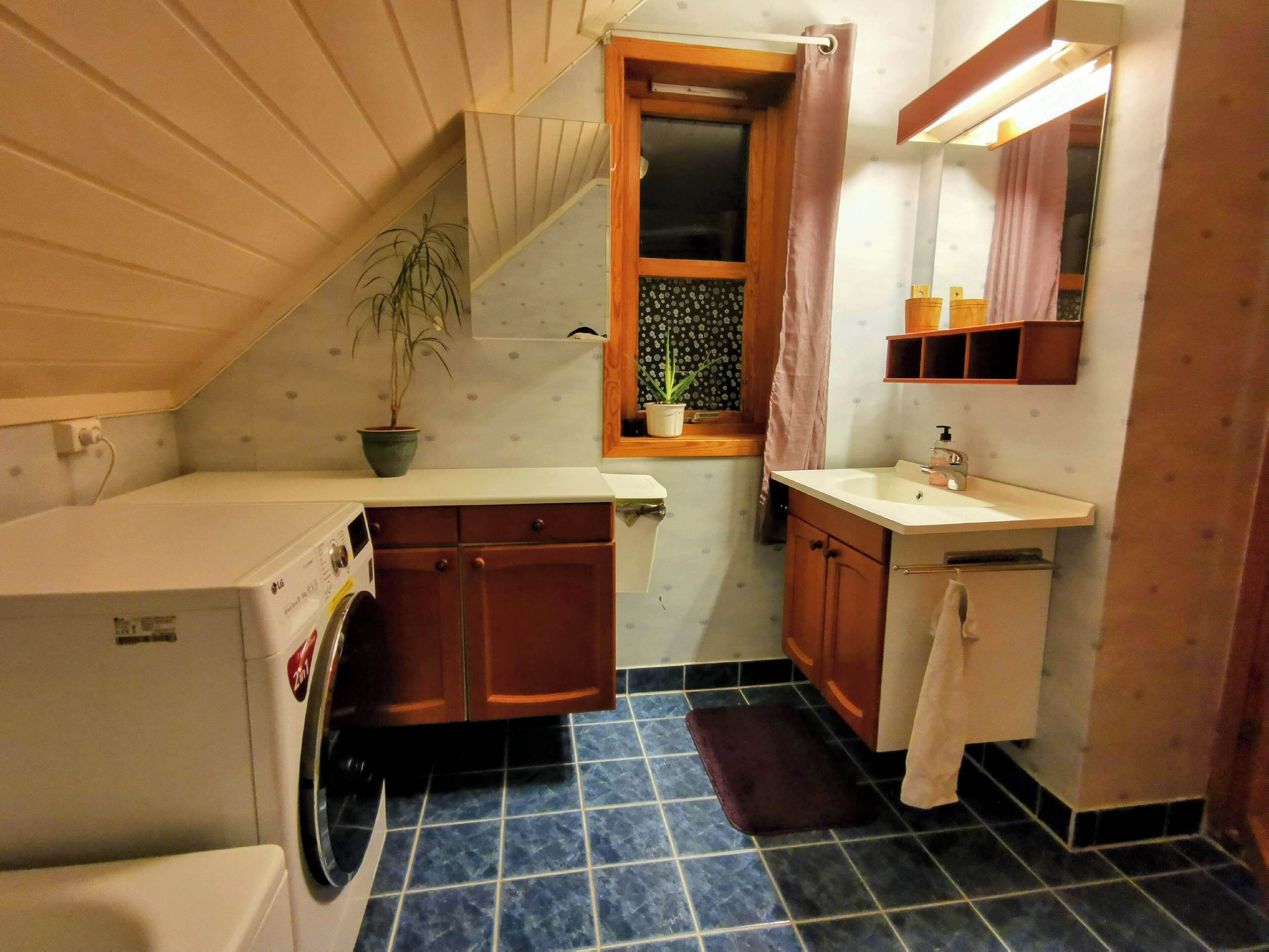 A compact kitchen with a washing machine, wooden cabinets, a small window with a curtain, and a sink with a mirror above it.