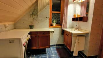 A compact kitchen with a washing machine, wooden cabinets, a small window with a curtain, and a sink with a mirror above it.