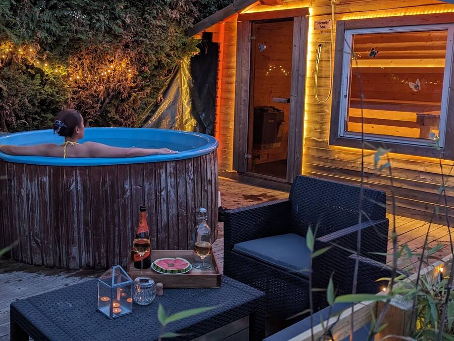 A wooden hot tub with a person relaxing, surrounded by outdoor furniture and a glass enclosure.