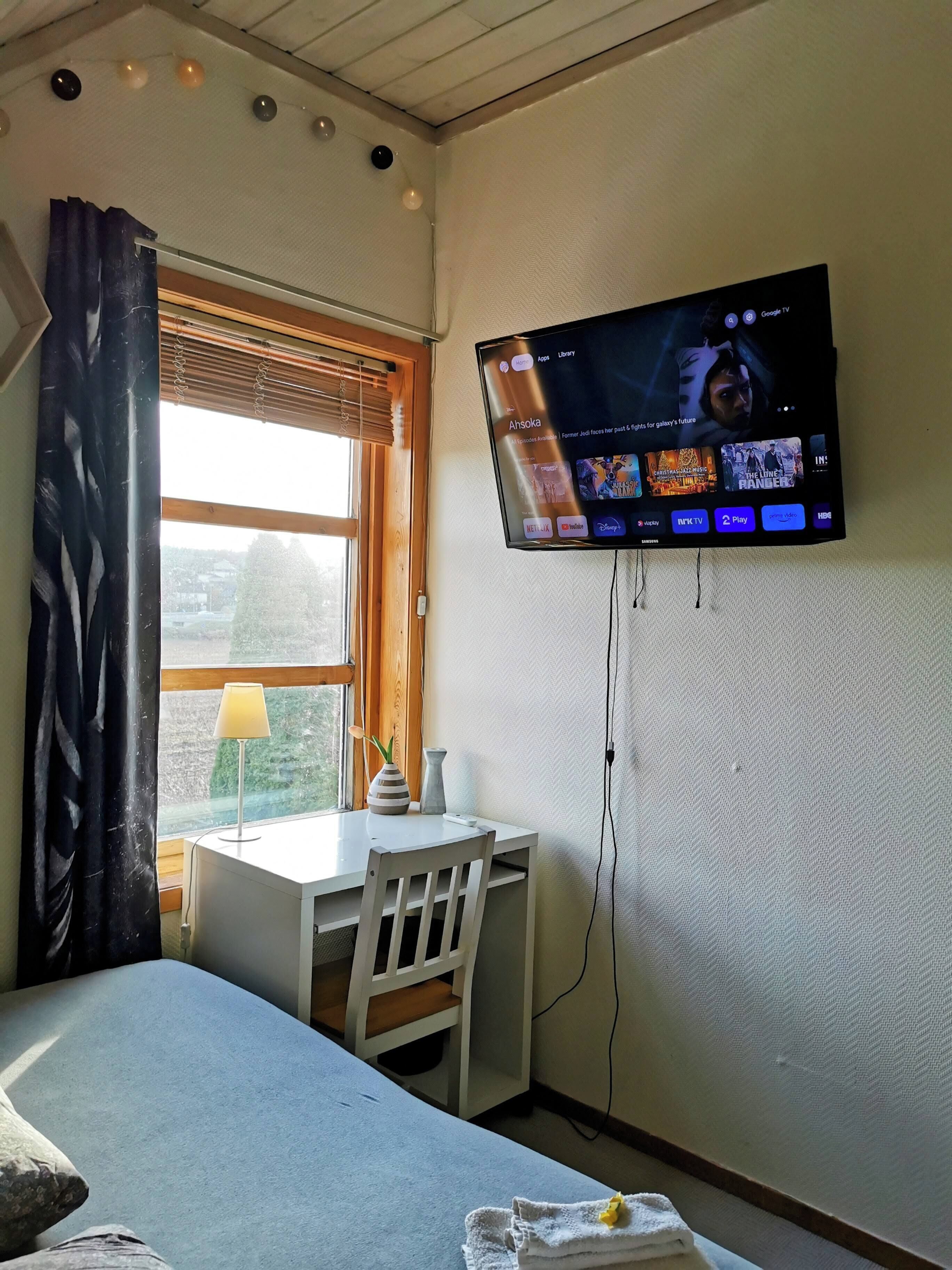 A hotel room with a bed, a desk with a lamp, a TV mounted on the wall displaying a streaming service interface, and a window with blinds.