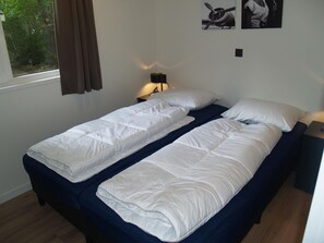 Cama doble con ropa de cama blanca en una habitación con ventana, lámpara y dos cuadros en la pared.