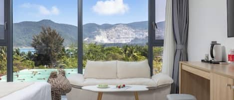 A room with a sofa, a small table, a desk, and a view of mountains and a beach.
