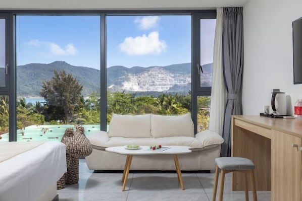 Quarto com sofá, mesa pequena, escrivaninha e vista para montanhas e praia.