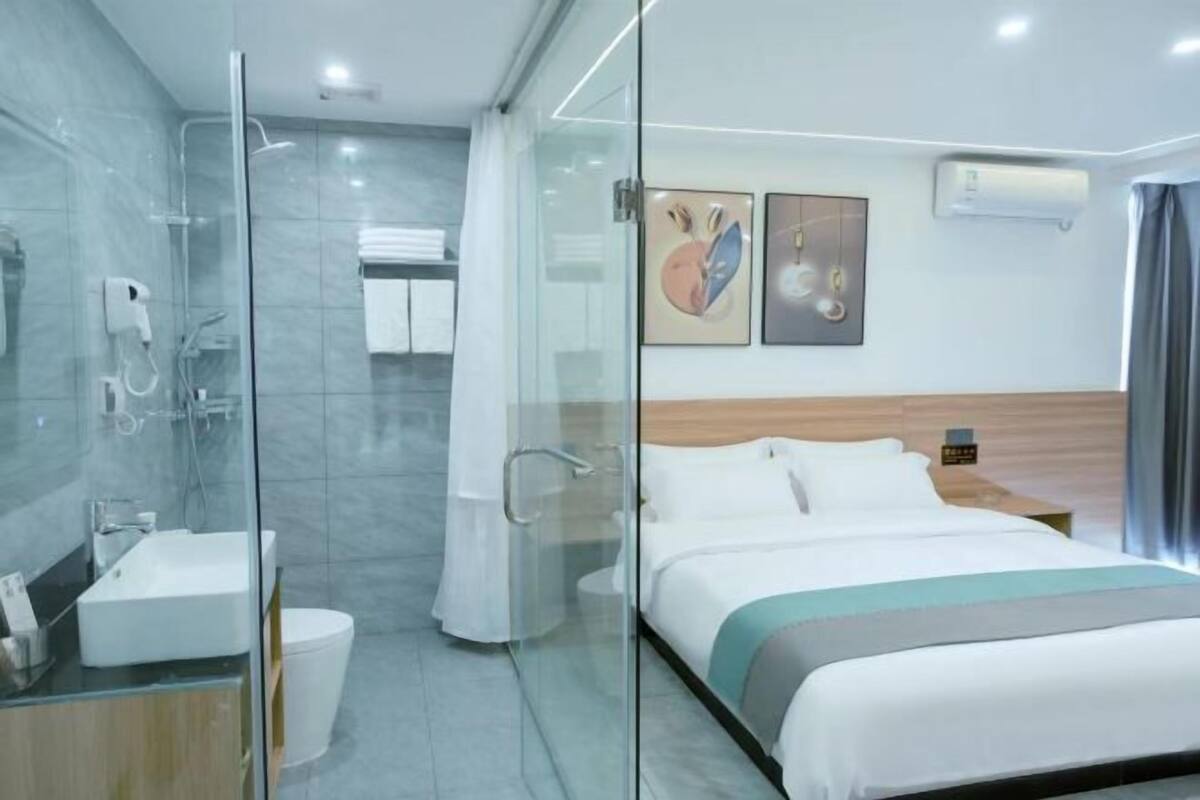 Une chambre d’hôtel moderne dotée d’une douche à parois de verre, d’un lit avec une literie blanche, d’une table de chevet et d’un climatiseur fixé au mur.